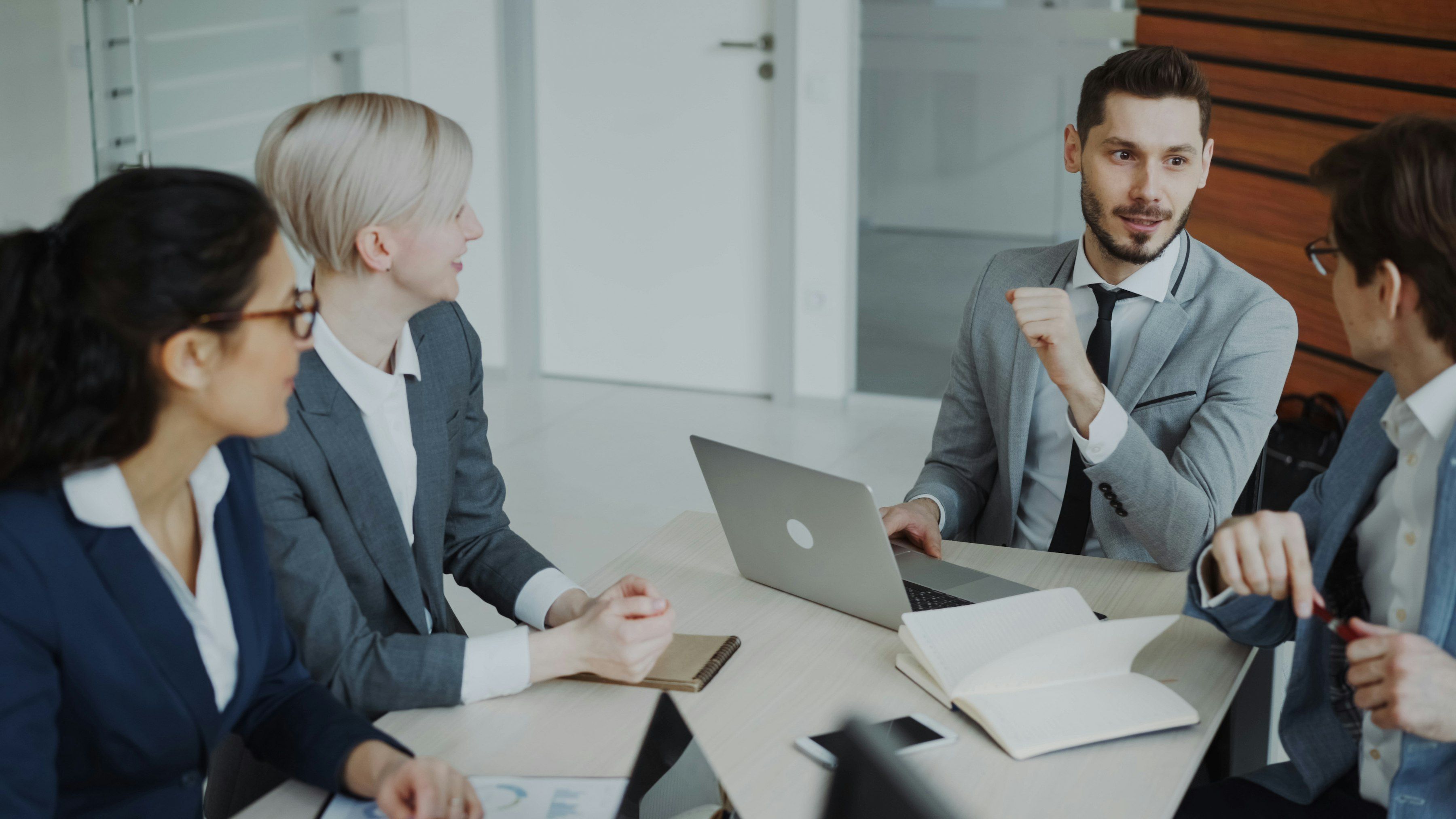 Entretien de conseil autour d’un ordinateur portable : un homme en costume gris explique un point à ses interlocutrices, carnet ouvert sur la table.